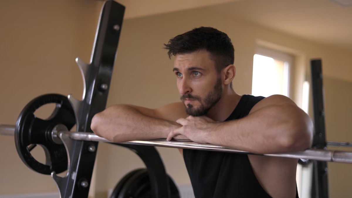 Concentrated muscular bearded sportsman in black activewear leaning on metal barbell and looking away while resting during training in modern gym